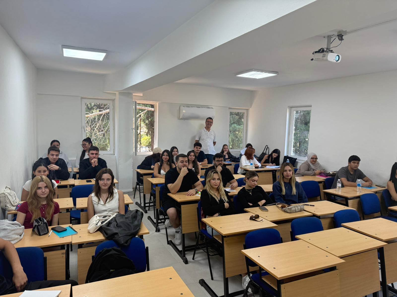 Dalaman MYO’da geniş ve aydınlık bir derslikte gerçekleştirilen toplantı. Sıralarda oturan kalabalık bir öğrenci grubu kameraya dönük halde poz veriyor. Arka tarafta ayakta duran bir öğretim elemanı görülüyor. Derslikte mavi sandalyeler, ahşap sıralar ve büyük pencereler bulunuyor.